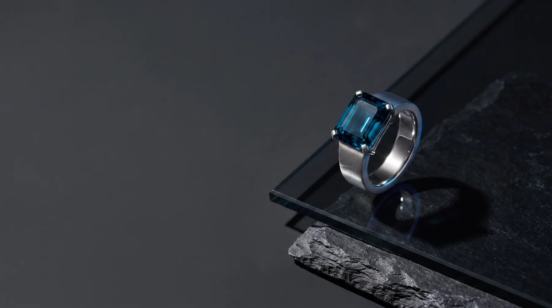 A deep teal-blue emerald-cut gemstone in a brushed platinum ring, photographed on smoked glass and dark slate.
