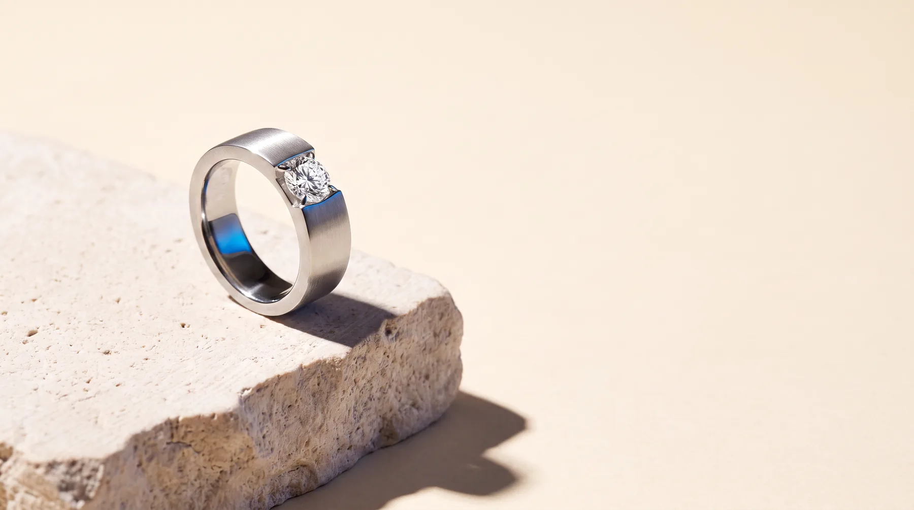 A brushed platinum ring set with a round brilliant diamond, photographed on a pale limestone block against a warm ivory background.