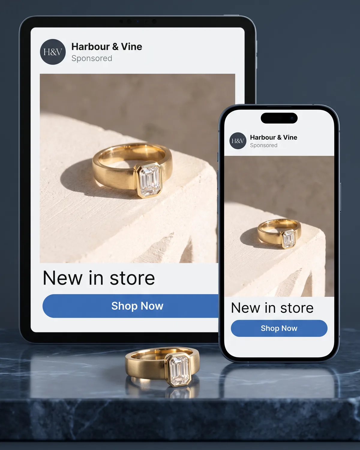 A tablet and a phone standing on a marble slab, both showing the same sample social media advertisement for a sample jewelry store, Harbour & Vine — a photograph of a yellow gold ring with an emerald-cut diamond above the words “New in store” and a Shop Now button — with that same ring resting on the marble in front of them.