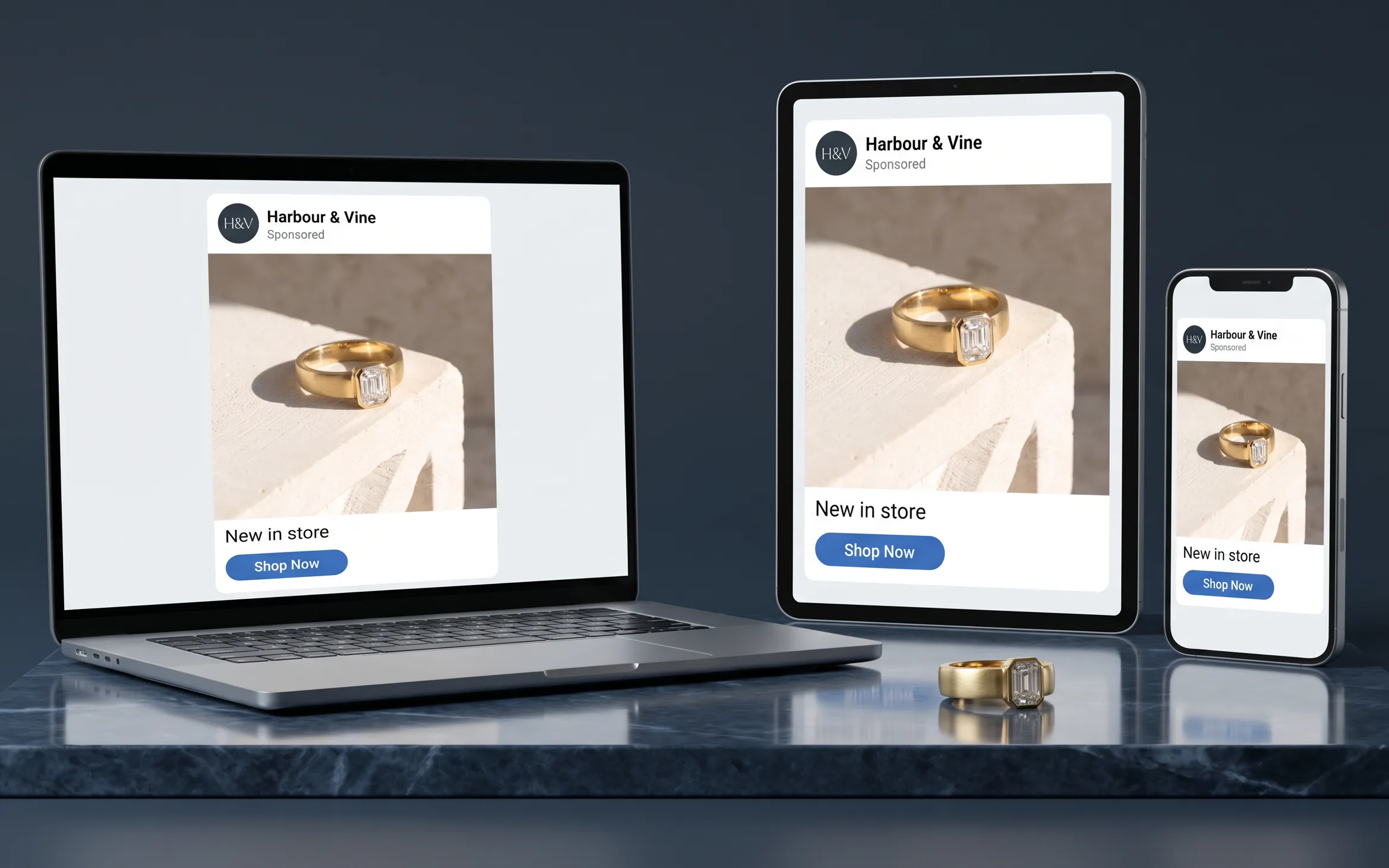 A laptop, tablet and phone standing together on a marble slab, each showing the same sample social media advertisement for a sample jewelry store, Harbour & Vine — a photograph of a yellow gold ring with an emerald-cut diamond above the words “New in store” and a Shop Now button — with that same ring resting on the marble in front of them.