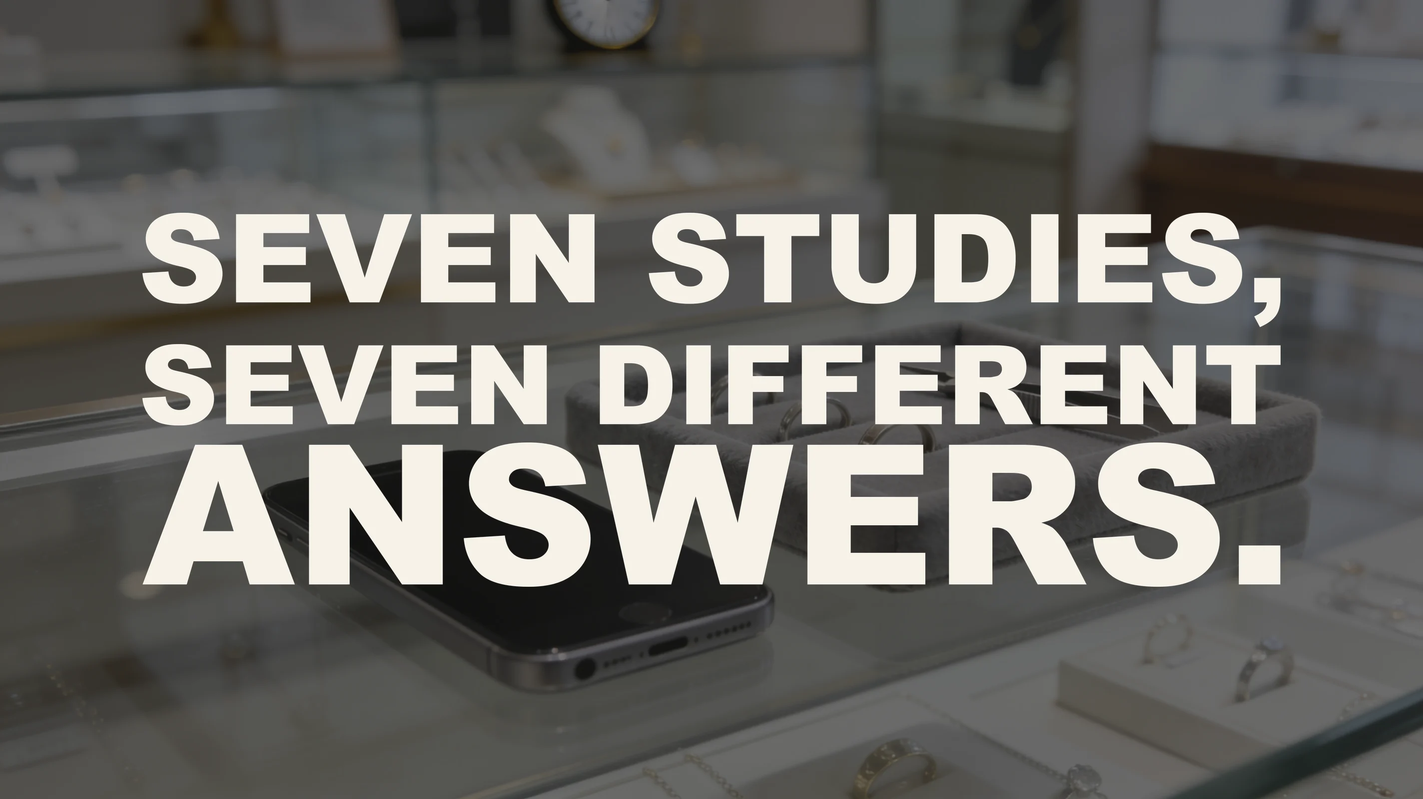 The words “Seven studies, seven different answers.” set in large type over a photograph taken on the glass counter of a jewelry store — a smartphone lies face up with its screen completely dark beside a grey suede ring tray holding plain bands and a pair of tweezers, with the shop’s display cases and a wall clock softly out of focus behind.
