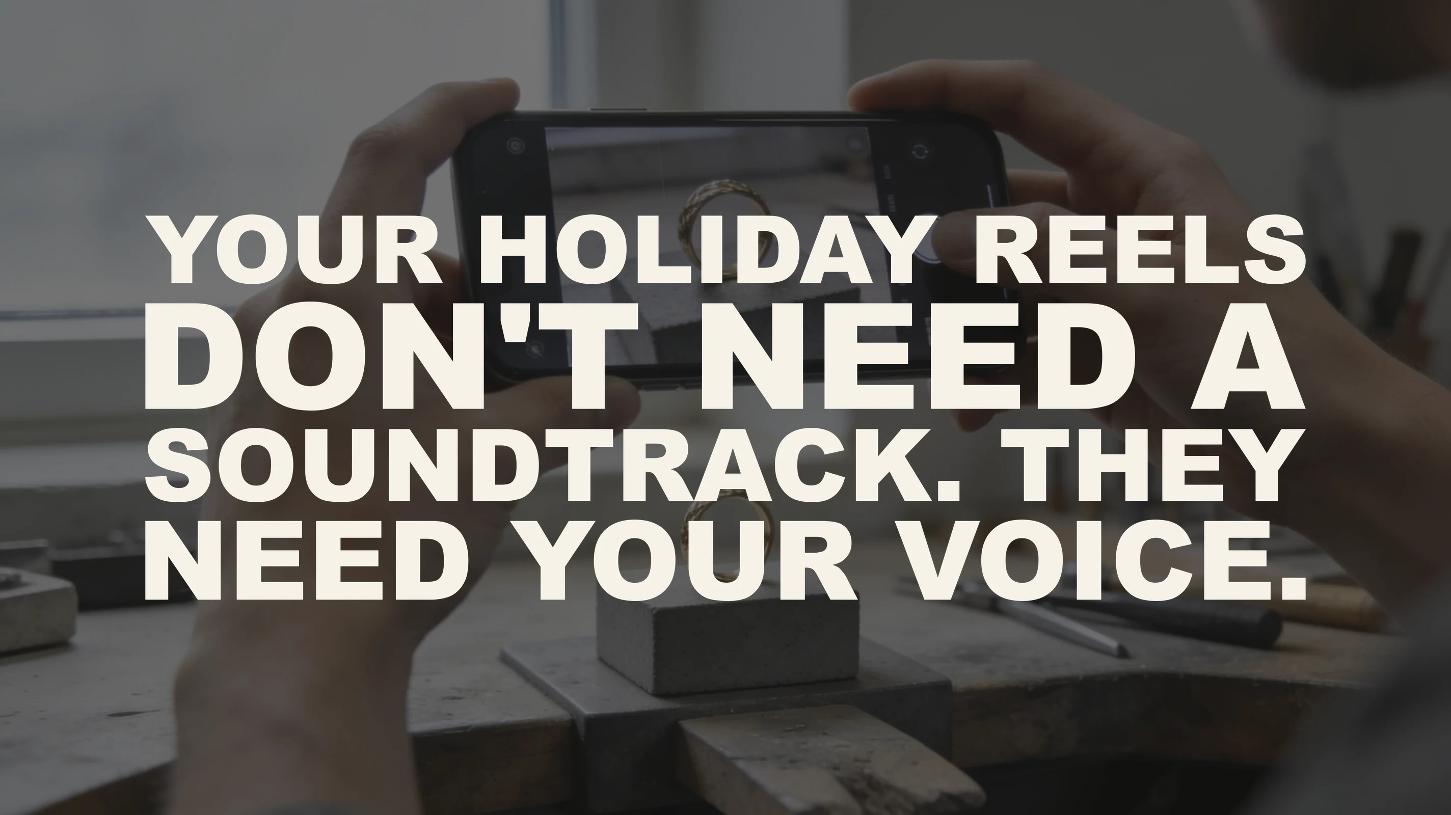 The words "Your holiday Reels don't need a soundtrack. They need your voice." set in large type over a photograph of a jeweler's hands filming a gold ring with a smartphone at the workbench, the ring visible both on the bench and on the phone's screen, in overcast window light.