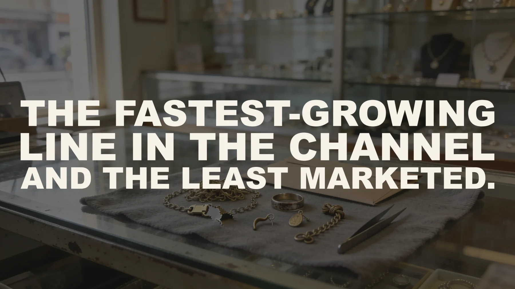 The words “The fastest-growing line in the channel and the least marketed.” set in large type over a photograph taken inside a jewelry store — a customer's own old gold laid out on a grey cloth on the glass counter: a tangled chain with a snapped clasp, a worn wedding band, one odd earring, a pendant and a knotted chain, beside a plain kraft job envelope and a pair of tweezers, with full display cases and daylight from the shop window behind.