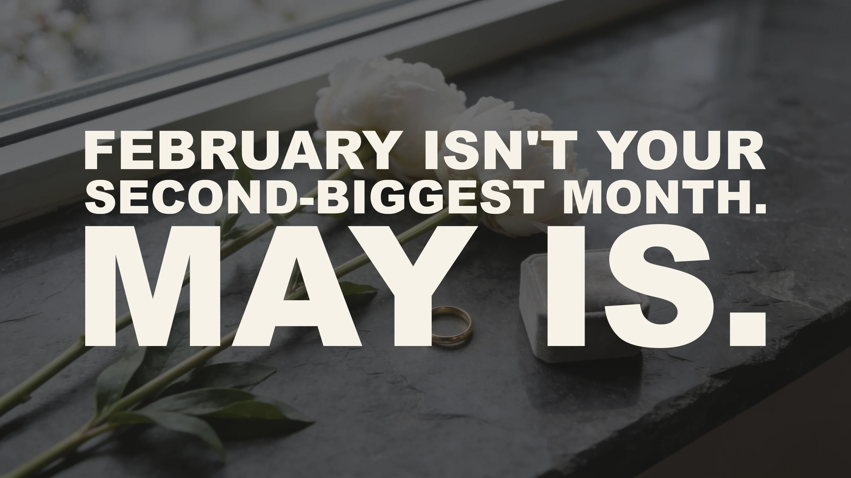 The words "February isn't your second-biggest month. May is." set in large type over a photograph of two white peonies lying on a graphite stone windowsill beside a plain gold band and a grey velvet ring box, in overcast spring daylight.