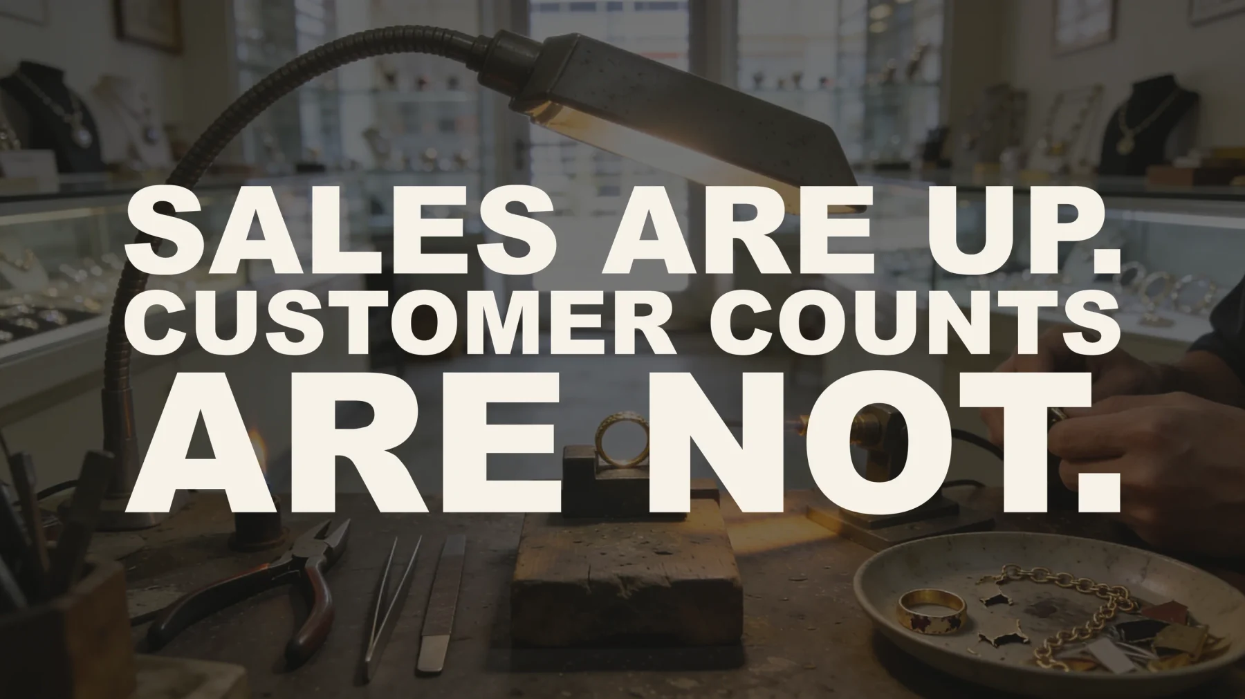The words “Sales are up. Customer counts are not.” set in large type over a photograph of a jeweler's bench — a gold ring held in a clamp under a torch flame, hand tools laid out, and a dish holding a customer's own broken chain and worn wedding band, with the store's full display cases and the front door beyond.
