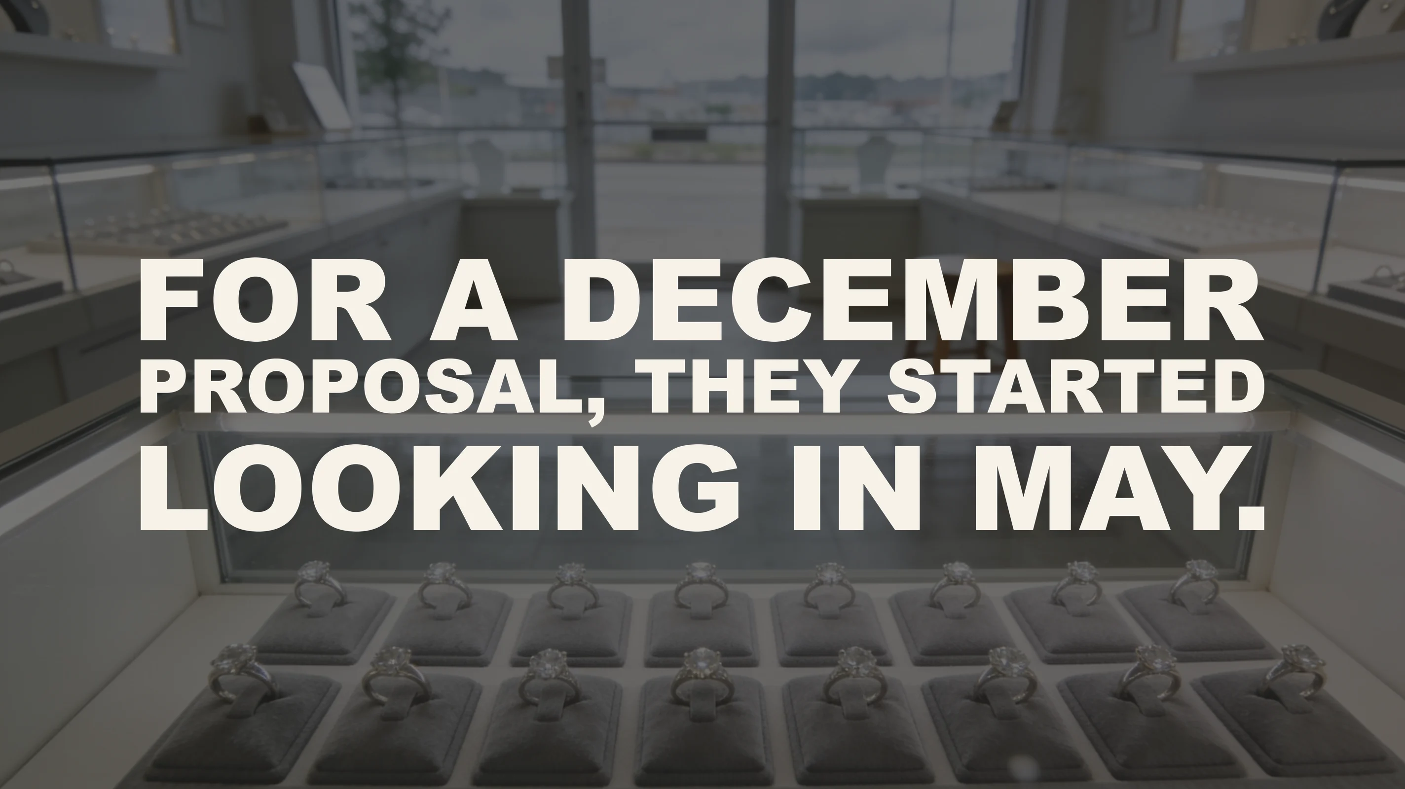 The words “For a December proposal, they started looking in May.” set in large type over a photograph taken inside an independent jewelry store with nobody in it — a lit case of solitaire engagement rings on grey suede pads fills the foreground, and behind it the empty shop floor recedes past more display cases and an unoccupied wooden stool to a glass door full of flat overcast daylight.