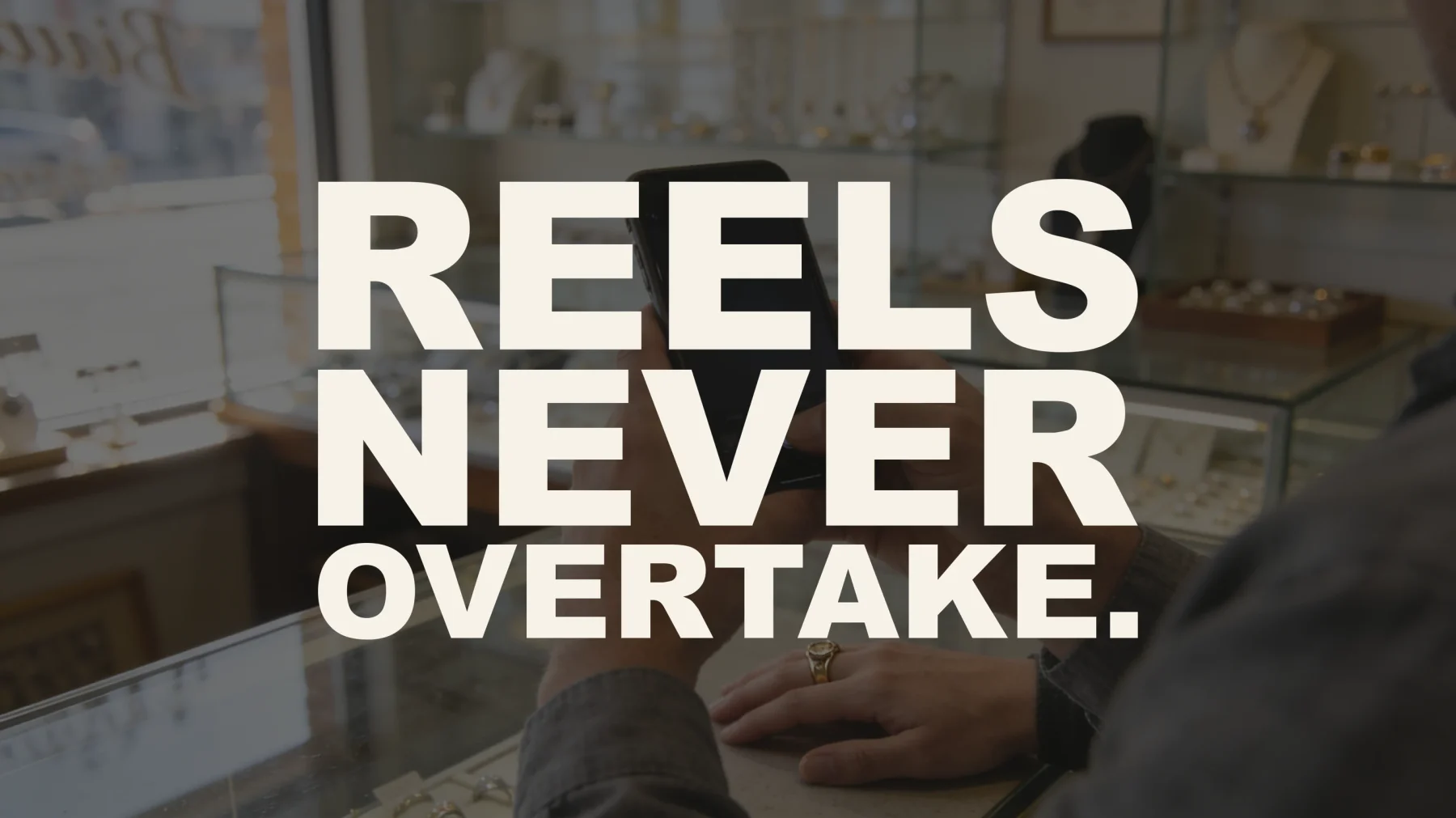 The words “Reels never overtake.” set in large type over a photograph taken inside a small jewelry store — a jeweler holding a phone to photograph a gold ring worn on a customer's hand resting on the glass counter, with display cases and daylight from the shop window behind.